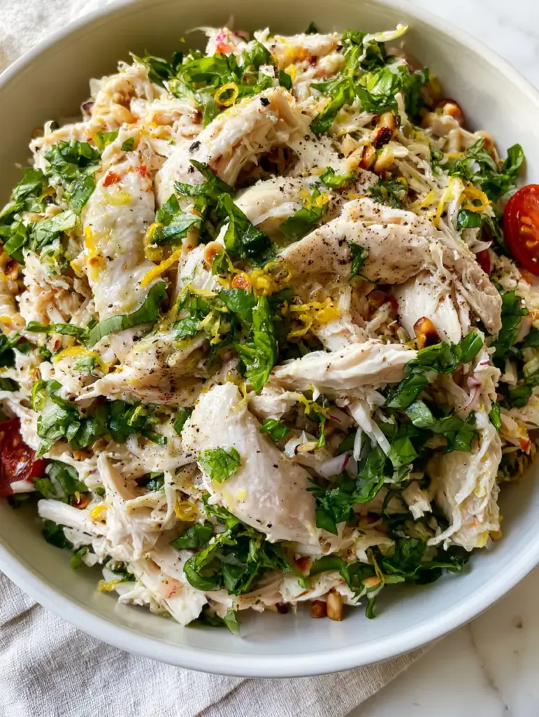 Lemon Basil Chicken Salad with Toasted Pine Nuts and Fresh Herbs 1 Lemon Basil Chicken Salad served in a white ceramic bowl on a marble countertop with halved cherry tomatoes and pine nuts.